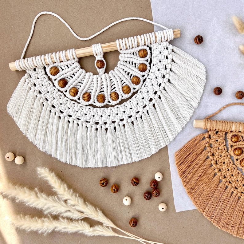 May include: A white macrame wall hanging with a wooden dowel and decorative wooden beads. The handcrafted piece features a fan-like design with intricate knotting and fringe. Additional wooden beads and decorative elements are scattered around the wall hanging.