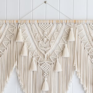 May include: Cream-colored macrame wall hanging with intricate geometric patterns and tassels. The handcrafted textile art piece features a wooden dowel for hanging and adds a bohemian touch to any space. The macrame design is symmetrical and detailed.