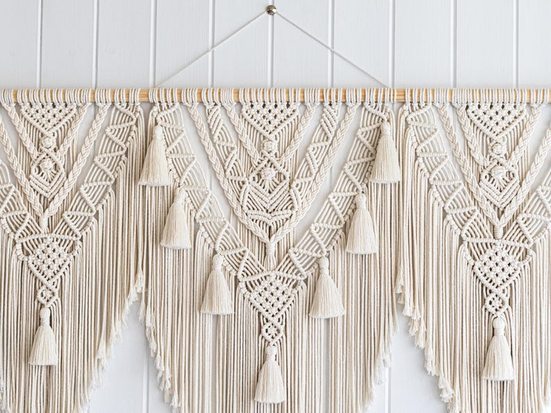 May include: Cream-colored macrame wall hanging with intricate geometric patterns and tassels. The handcrafted textile art piece features a wooden dowel for hanging and adds a bohemian touch to any space. The macrame design is symmetrical and detailed.