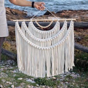 Puede incluir: Colgante de pared de macramé color crema con una clavija de madera y detalles de cuentas. La pieza artesanal presenta múltiples capas de cordón de algodón anudado, creando un diseño bohemio texturizado. La decoración de la pared está sostenida por una mano.