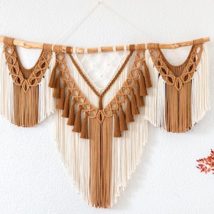 Op de afbeelding: Een macramé wandkleed in bruin- en crèmetinten, opgehangen aan een natuurlijke houten deuvel. Het ontwerp heeft ingewikkelde knoopmotieven, kwasten en een geometrische vorm, wat een bohemien touch toevoegt aan de woondecoratie.