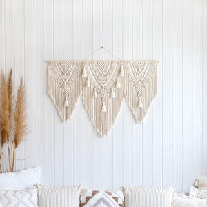May include: A large, cream-colored macrame wall hanging with a geometric design and tassels. The wall decor is mounted on a wooden dowel and hangs against a white shiplap wall. Decorative pillows and dried plants are in the foreground.