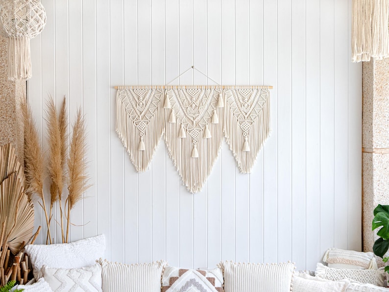 May include: A large, cream-colored macrame wall hanging with a geometric design and tassels. The wall decor is mounted on a wooden dowel and hangs against a white shiplap wall. Decorative pillows and dried plants are in the foreground.