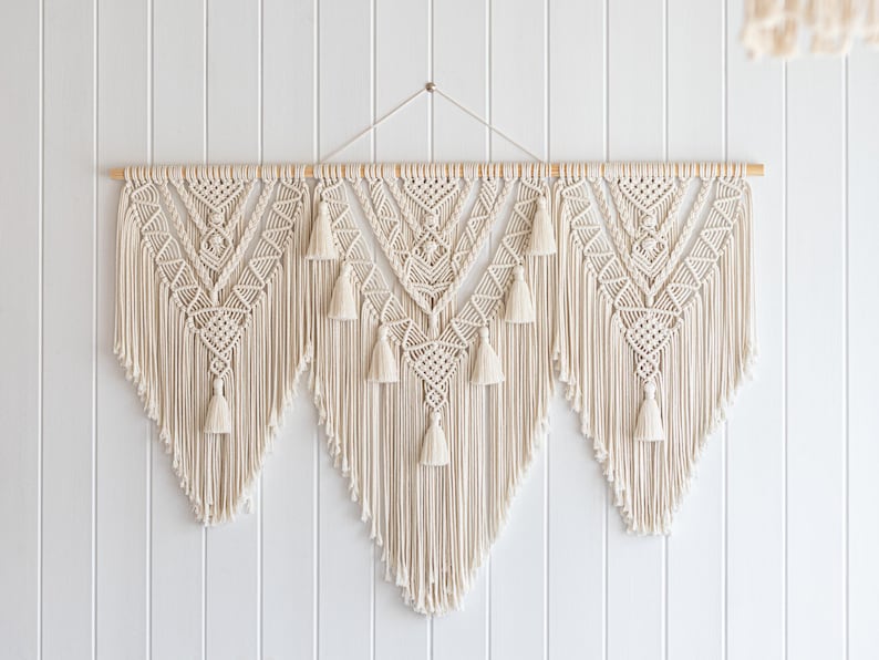 May include: Cream-colored macrame wall hanging with geometric patterns and tassels. The handcrafted textile art piece is suspended from a wooden dowel, adding a bohemian touch to home decor. The wall hanging is approximately 40 inches wide.