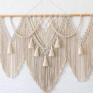 Könnte beinhalten: Cremefarbener Makramee-Wandbehang mit einem Holzstab. Das aufwendige Design weist geometrische Muster, Quasten und einen kaskadierenden Fransenbesatz auf. Diese Dekoration im Bohème-Stil verleiht jedem Raum Textur und Wärme.