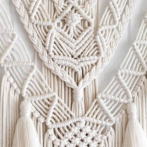 May include: Cream-colored macrame wall hanging with intricate geometric patterns and braided edges. The design features diamond shapes and tassels, creating a textured, bohemian aesthetic. This handcrafted piece adds a touch of natural elegance to any space.