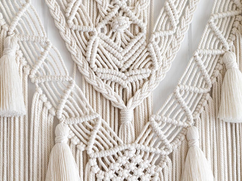 May include: Cream-colored macrame wall hanging with intricate geometric patterns and braided edges. The design features diamond shapes and tassels, creating a textured, bohemian aesthetic. This handcrafted piece adds a touch of natural elegance to any space.