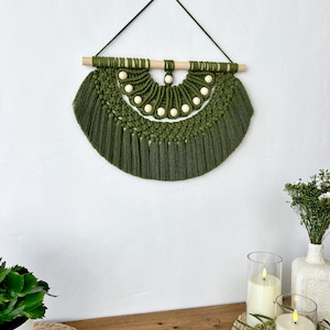 May include: A handmade olive green macrame wall hanging. The wall decor features a half-circle design with a wooden dowel and small wooden beads. The macrame has a fringe at the bottom and is suspended by a dark green cord.