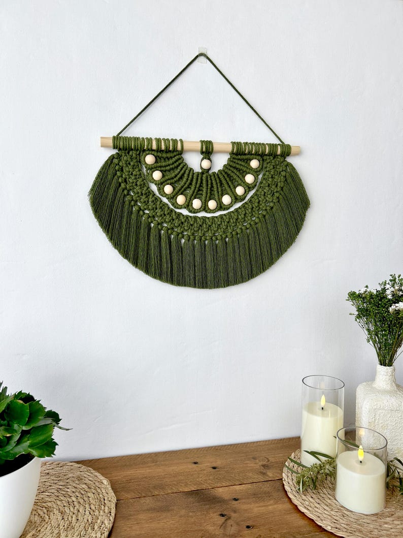 May include: A handmade olive green macrame wall hanging. The wall decor features a half-circle design with a wooden dowel and small wooden beads. The macrame has a fringe at the bottom and is suspended by a dark green cord.