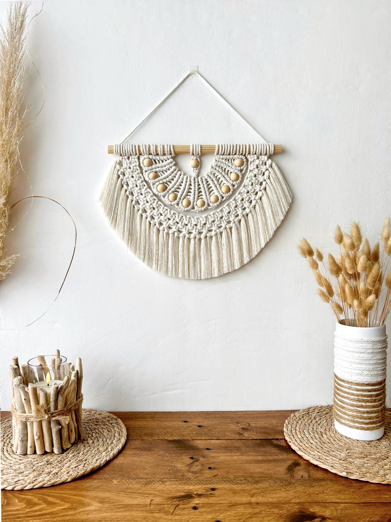 May include: A cream-colored macrame wall hanging with a half-circle design. The piece features a wooden dowel at the top, wooden beads, and knotted fringe. The wall hanging is displayed against a white wall, with decorative elements on a wooden table.