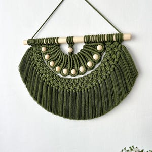 May include: Olive green macrame wall hanging with a wooden dowel and natural wooden beads. The handcrafted piece features a half-circle design with a fringe at the bottom. A green cord is used for hanging.