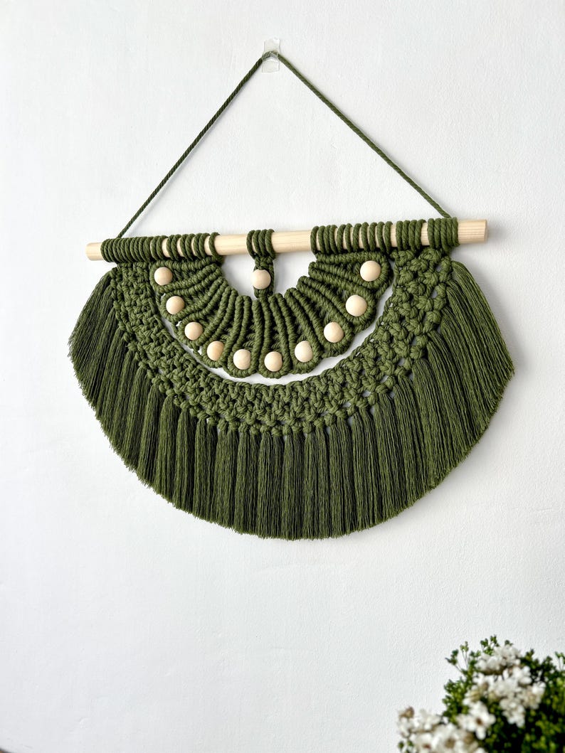 May include: Olive green macrame wall hanging with a wooden dowel and natural wooden beads. The handcrafted piece features a half-circle design with a fringe at the bottom. A green cord is used for hanging.