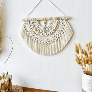 May include: Cream-colored macrame wall hanging with a half-circle design. The piece features a wooden dowel at the top, adorned with wooden beads. The macrame has a textured pattern with fringe at the bottom, creating a bohemian aesthetic.