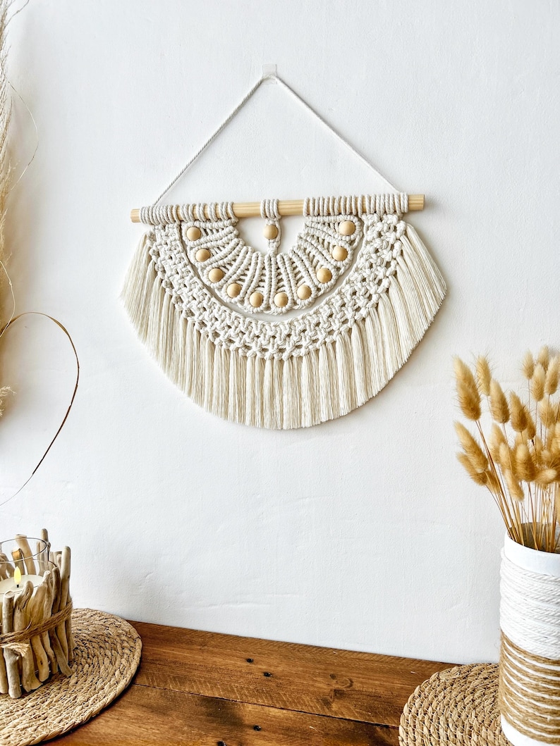 May include: Cream-colored macrame wall hanging with a half-circle design. The piece features a wooden dowel at the top, adorned with wooden beads. The macrame has a textured pattern with fringe at the bottom, creating a bohemian aesthetic.