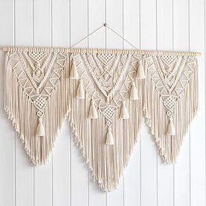 May include: Cream-colored macrame wall hanging with a wooden dowel. The design features geometric patterns and tassels, creating a bohemian aesthetic. This handcrafted piece adds texture and visual interest to any space.