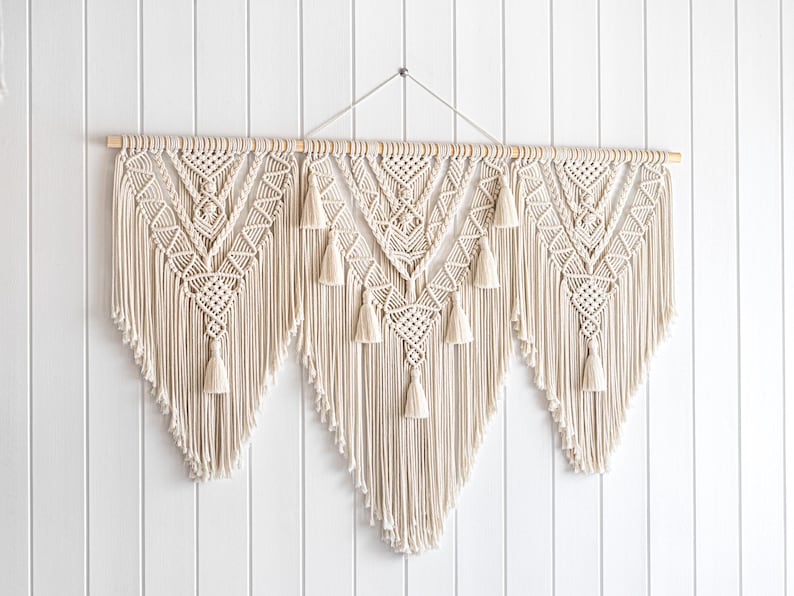 May include: Cream-colored macrame wall hanging with a wooden dowel. The design features geometric patterns and tassels, creating a bohemian aesthetic. This handcrafted piece adds texture and visual interest to any space.