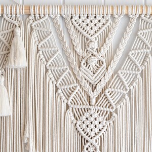 May include: Cream-colored macrame wall hanging with intricate geometric patterns and knotted details. Features a wooden dowel for hanging and decorative tassels. A handcrafted, bohemian-style home decor item.