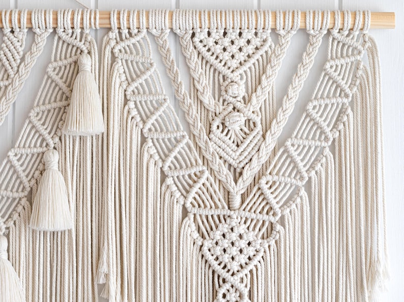 May include: Cream-colored macrame wall hanging with intricate geometric patterns and knotted details. Features a wooden dowel for hanging and decorative tassels. A handcrafted, bohemian-style home decor item.