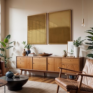 A warm, sunlit living room with mid-century furniture, tan leather seating, and lush indoor plants. A wooden credenza anchors the space beneath the Amber & Dawn Divide Set. The two minimalist artworks feature soft amber and golden brown gradients on linen, blending perfectly with the natural textures and earthy palette, creating a sense of quiet balance and modern organic harmony.