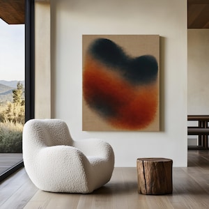 May include: An abstract painting with a blurred design in shades of black, orange, and red hangs on a beige wall. A white, fluffy armchair sits in front of a large window. A small, wooden stump serves as a side table.
