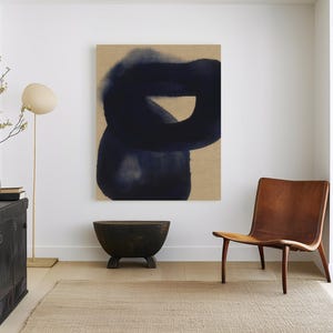 May include: A large rectangular wall art piece featuring a dark navy blue abstract ink-wash shape on a tan background, displayed in a room with a brown leather chair, a black cabinet, a small dark bowl, and a floor lamp with a beige shade.