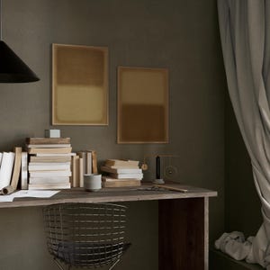 A cozy, moody workspace with stacked books, a rustic wooden desk, and soft draped curtains. Above the desk hang two framed minimalist prints the Amber & Dawn Divide Set, featuring soft ochre and golden brown tones that blend into natural linen. The warm abstract art adds balance and quiet sophistication to the intimate study space, complementing the earthy wall and subdued lighting.