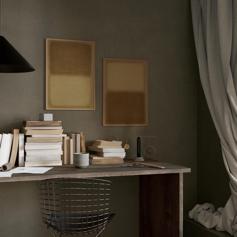 A cozy, moody workspace with stacked books, a rustic wooden desk, and soft draped curtains. Above the desk hang two framed minimalist prints the Amber & Dawn Divide Set, featuring soft ochre and golden brown tones that blend into natural linen. The warm abstract art adds balance and quiet sophistication to the intimate study space, complementing the earthy wall and subdued lighting.