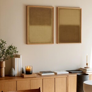 A cozy, moody workspace with stacked books, and a wooden desk. Above the desk hang two framed minimalist prints the Amber & Dawn Divide Set, featuring soft ochre and golden brown tones that blend into natural linen. The warm abstract art adds balance and quiet sophistication to the intimate study space, complementing the earthy wall and subdued lighting.