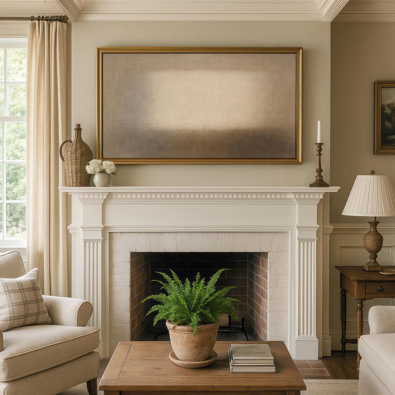 A warm living room with cream walls, classic molding, a white mantel fireplace, and neutral furnishings is decorated with warm lighting, and a potted fern centered on a wooden coffee table. Displayed above the mantel on a Frame TV is soft beige and earth-toned layers with a diffused central form and subtle textured surface. Designed for Frame TV, this neutral artwork creates a calm, gallery-style presence on a television, blending naturally into modern, Japandi, and minimalist interiors.