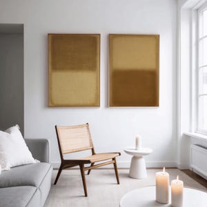 A bright, minimalist living room with a soft gray sofa, woven accent chair, and white tables topped with candles. On the wall, two framed abstract prints, Amber & Dawn Divide Set—glow in warm ochre and golden tones on natural linen. The diptych brings balance, calm, and understated elegance to the serene Japandi-style space filled with natural light.