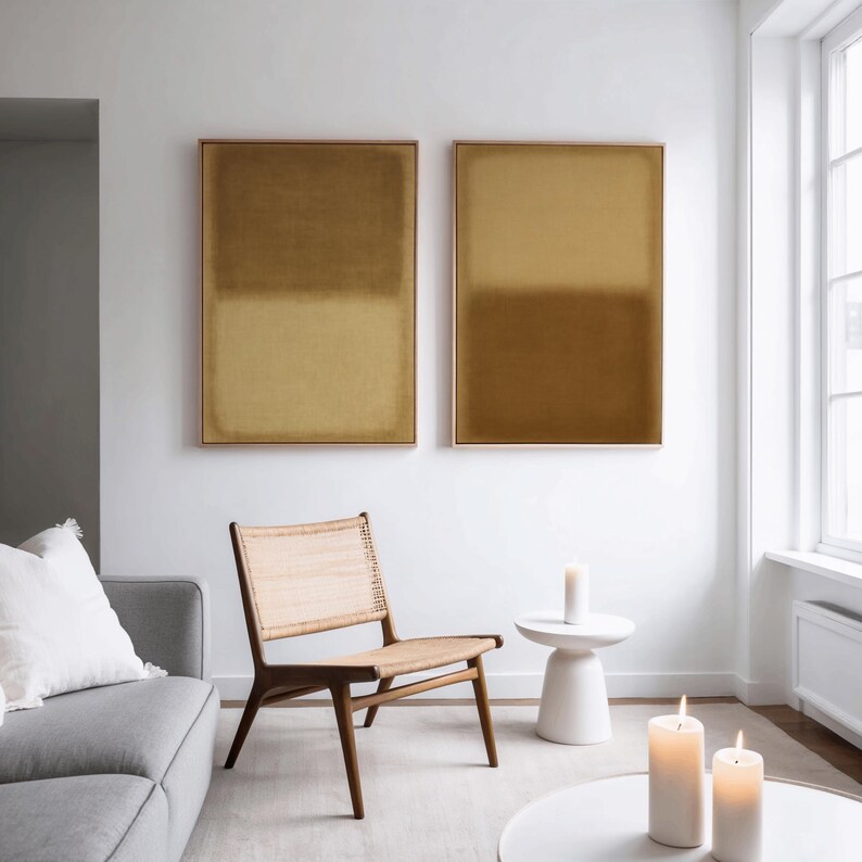A bright, minimalist living room with a soft gray sofa, woven accent chair, and white tables topped with candles. On the wall, two framed abstract prints, Amber & Dawn Divide Set—glow in warm ochre and golden tones on natural linen. The diptych brings balance, calm, and understated elegance to the serene Japandi-style space filled with natural light.