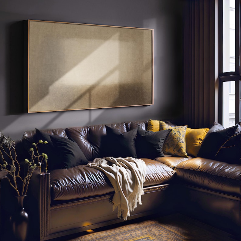 A moody living room features dark walls, a deep brown leather sectional, and mustard yellow accent pillows, creating a dramatic and cozy atmosphere. On the wall above, the Samsung Frame TV displays soft beige and earth-toned layers with a diffused central form and subtle textured surface. Designed for Frame TV, this neutral artwork creates a calm, gallery-style presence on a television, blending naturally into modern, Japandi, and minimalist interiors.