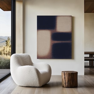 Puede incluir: Una pintura abstracta con una combinación de colores azul marino y beige cuelga de una pared blanca. Un sillón blanco y esponjoso se encuentra frente a una gran ventana. Un pequeño tocón de árbol de madera sirve como mesa auxiliar.