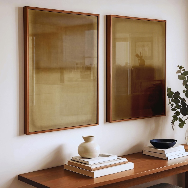A warm, modern interior with a wooden console table styled with stacked art books, ceramic vases, and fresh greenery. Above the table hang two framed minimalist prints, the Amber & Dawn Divide Set featuring layered ochre tones and linen texture. Soft sunlight enhances the natural hues and organic calm of the diptych, creating a serene, Japandi-inspired atmosphere.