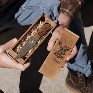 May include: A folding pocket knife with a wooden handle and a patterned blade, presented in a wooden box with decorative straw. The box lid is engraved with a deer design and the name "JACKSON" and the date "07.20.25".
