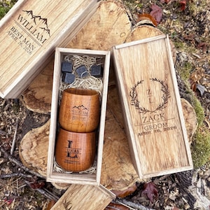 May include: Wooden gift set featuring two engraved wooden tumblers and whiskey stones, nestled in a wooden box. The box and tumblers are engraved with names, titles, and dates. The set is presented on a natural wood surface.