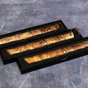 May include: Three wooden knife cases with personalized names, "WZ", "JD", and "William", engraved on the wood. The cases are black and have a leather strap.