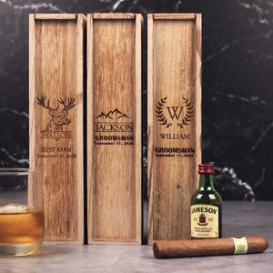 May include: Three wooden gift boxes with engraved names and titles, including "Watson, Best Man," "Jackson, Groomsman," and "William, Groomsman," with a date. A miniature Jameson bottle and a cigar are also present.