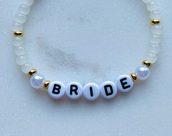 May include: White beaded bracelet with gold beads and white letter beads spelling out the word "BRIDE".