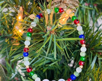 May include: A beaded bracelet with the word "JOY" spelled out in white letter beads. The bracelet features a mix of red, green, blue, and white beads, interspersed with gold-colored beads. The bracelet is displayed on a Christmas tree.