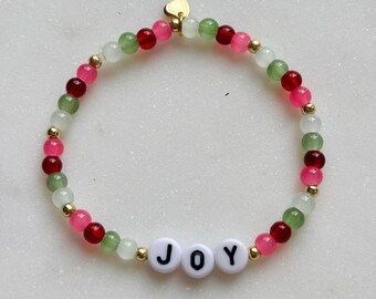 May include: A beaded bracelet with the word "JOY" spelled out in white letter beads. The bracelet features a mix of red, pink, green, and gold beads. A small gold heart charm is attached.