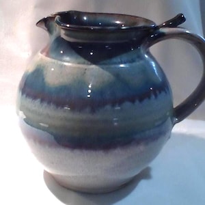 Może przedstawiać: Ręcznie wykonany ceramiczny dzbanek o zaokrąglonym korpusie i zakrzywionym uchwycie. Dzbanek ma paletę kolorów niebieskiego, fioletowego i białego, z błyszczącym wykończeniem. Wylewka jest przeznaczona do łatwego nalewania.