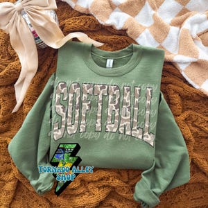 May include: An olive green sweatshirt with the word "SOFTBALL" in a camouflage pattern across the chest. The sweatshirt has a crew neck and long sleeves. A logo for "Tornado Alley Shop" is visible on the lower left.
