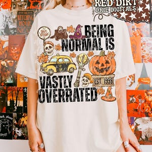 May include: A cream-colored t-shirt featuring a graphic print with the text "BEING NORMAL IS VASTLY OVERRATED" and "EST. 1998". The design includes a yellow taxi, a jack-o'-lantern, skulls, a witch hat, a bus stop sign, and autumn leaves.