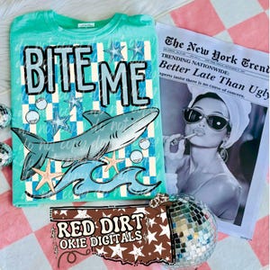 Può includere: Una maglietta turchese con la scritta "BITE ME", una grafica di squalo e il testo "RED DIRT OKIE DIGITALS". È visibile anche un giornale con il titolo "Better Late Than Ugly".
