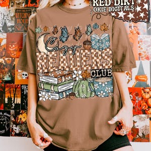 Op de afbeelding: Een bruin t-shirt met korte mouwen en een grafische print met de tekst "Cozy Readers Club". Het ontwerp bevat een halve maan, vlinders, een stapel boeken, een groene pompoen, een mok met bloemen en herfstbladeren in blauwe en oranje tinten.