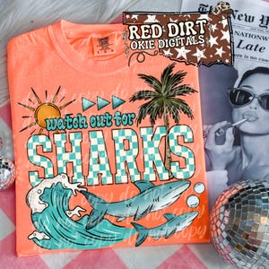 May include: Coral-colored t-shirt with a graphic design featuring a sun, palm tree, and sharks. The text "watch out for SHARKS" is prominently displayed in a checkered pattern. The shirt is a Comfort Colors brand.