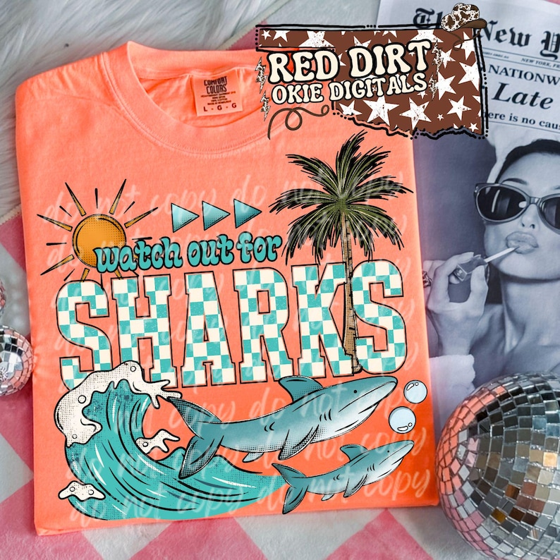 May include: Coral-colored t-shirt with a graphic design featuring a sun, palm tree, and sharks. The text "watch out for SHARKS" is prominently displayed in a checkered pattern. The shirt is a Comfort Colors brand.