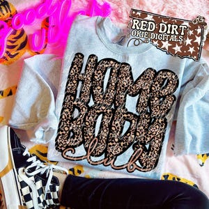 May include: A light blue sweatshirt with the text "Home Body Club" in a leopard print design. The sweatshirt is folded on a patterned surface, with a pair of black and white checkered sneakers visible.