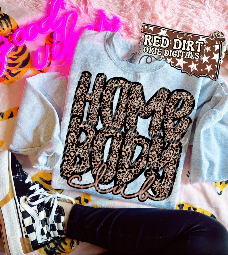 May include: A light blue sweatshirt with the text "Home Body Club" in a leopard print design. The sweatshirt is folded on a patterned surface, with a pair of black and white checkered sneakers visible.